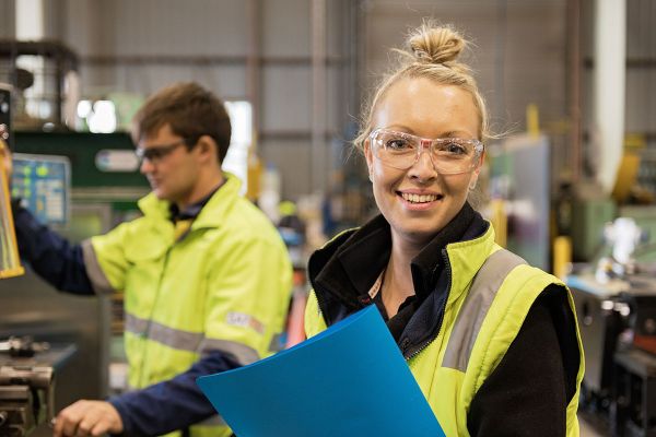 In a workshop, a young woman with blonde hair tied up in a bun, wearing clear protective glasses is holding a blue folder. Behind her is a man in protective clothing operating a machine.